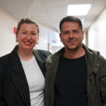 Two adults stand side by side in a hallway with white tiled walls and overhead fluorescent lighting, both looking at the camera and smiling.