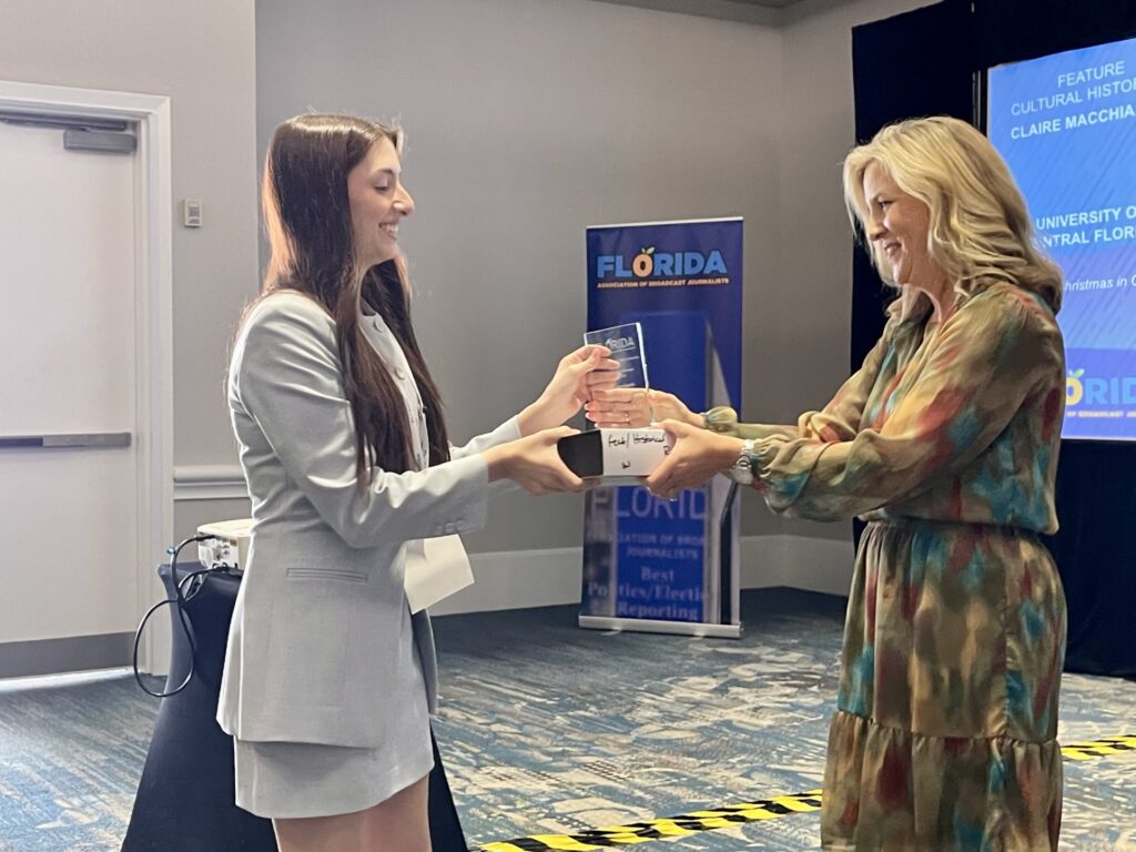 Two women stand indoors, smiling as one hands an award to the other. A "Florida" banner and presentation screen are visible in the background.