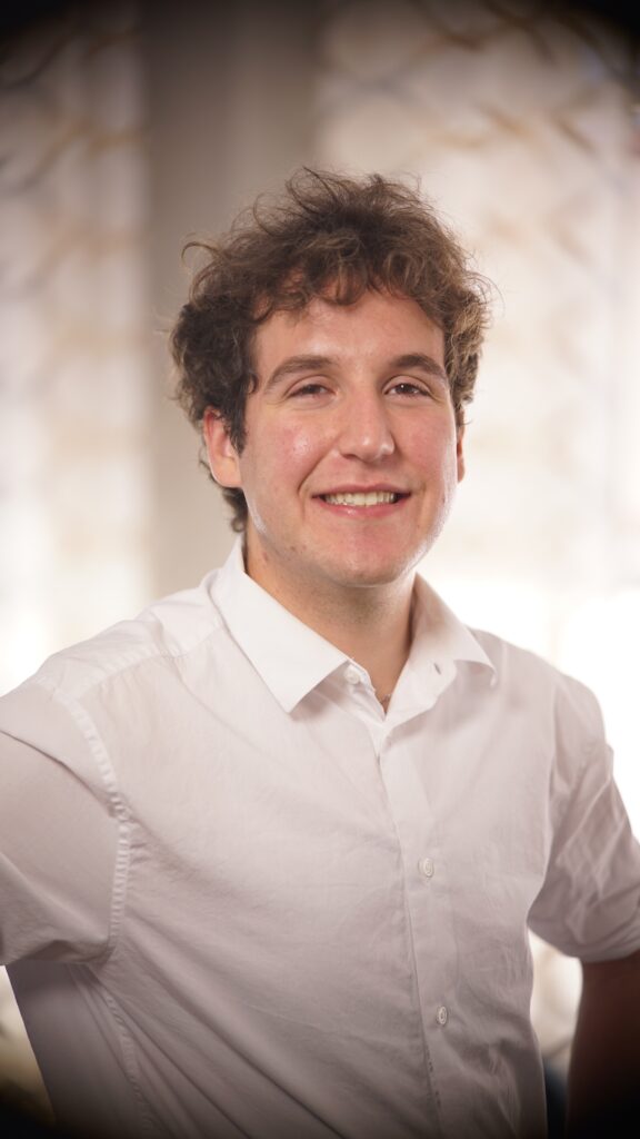 A young man with curly hair in a white button-up shirt stands indoors, smiling at the camera with a blurred background.
