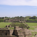 View of Monte Albán archaeological site with stone terraces, pyramids, and grassy plazas under a clear sky, with a few people walking among the ruins.