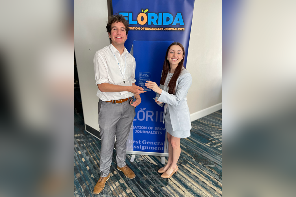 Two people stand in front of a "Florida Association of Broadcast Journalists" banner, holding an award together and smiling at the camera.