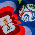 A FIFA World Cup soccer ball rests on a colorful surface featuring the FIFA logo and World Cup trophy graphic.