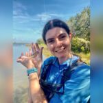 A person in a blue shirt stands outdoors by the water, smiling and holding a small fish in their hand on a sunny day.