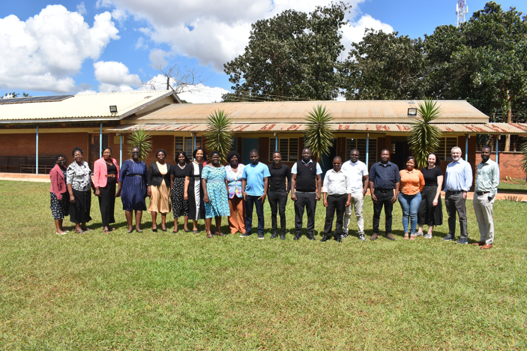 A group of 17 people stands in a line on grass in front of a building with trees and a partly cloudy sky in the background.