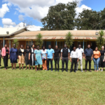 A group of 17 people stands in a line on grass in front of a building with trees and a partly cloudy sky in the background.