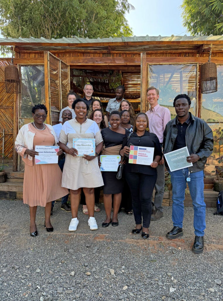 A group of people stand outside a wooden building, smiling and holding certificates, with greenery in the background.