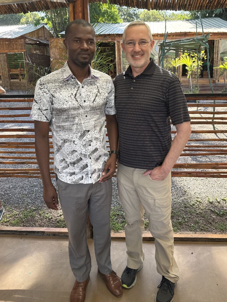 Two men stand side by side outdoors, smiling at the camera. Both are casually dressed, with a wooden fence and greenery in the background.
