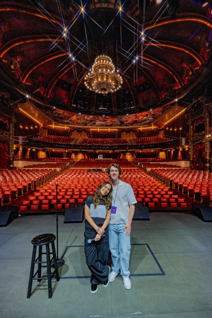 Two people stand on a theater stage with a stool and microphone in front of an empty, ornate auditorium featuring red seats and a large chandelier.