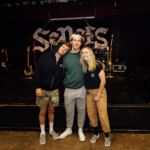 Three people stand together smiling in front of a stage with musical equipment and a large band logo in the background.
