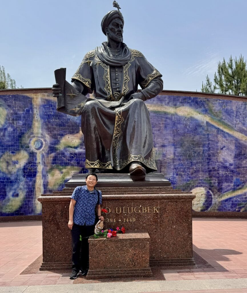A person stands beside a large statue of Ulugbek, holding a scroll, with flowers placed at the statue’s base and a blue mosaic wall in the background.