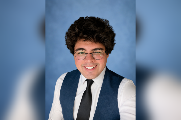 A person with curly brown hair, glasses, and a blue vest over a white shirt and black tie smiles at the camera against a plain blue background.
