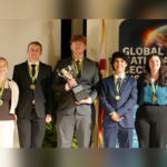 Five individuals wearing medals stand in a row, with one person holding a trophy. A 