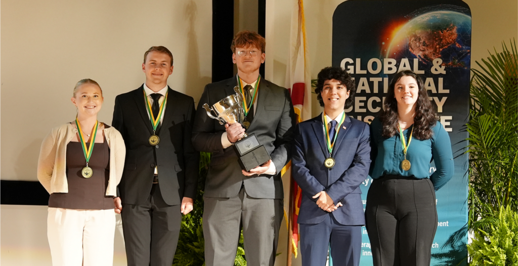 Five people wearing medals stand together; one holds a trophy. They are posed in front of a banner reading "Global & National Security Institute.