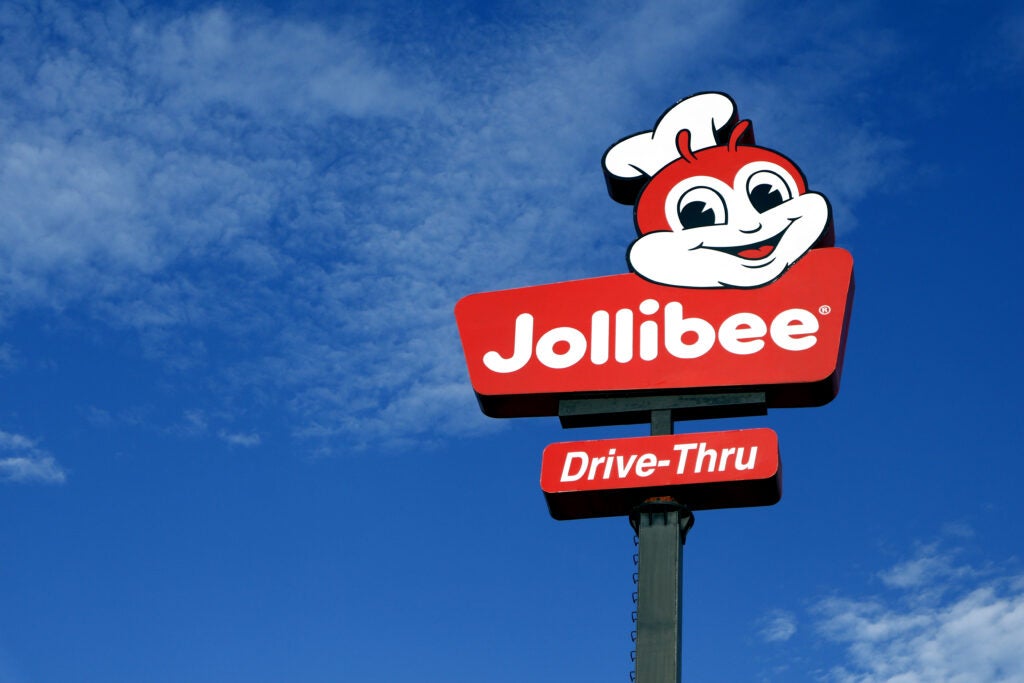 Jollibee drive-thru sign with mascot and logo against a blue sky with scattered clouds.
