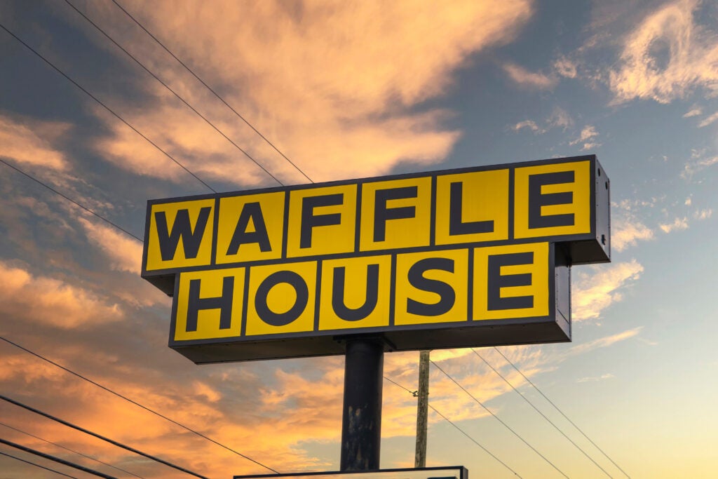 Waffle House sign with bold yellow and black lettering stands against a sunset sky with clouds and visible power lines.