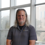 A person with long gray hair and a gray beard stands indoors by large windows, wearing a gray Laszlo Fityus Institute polo shirt.