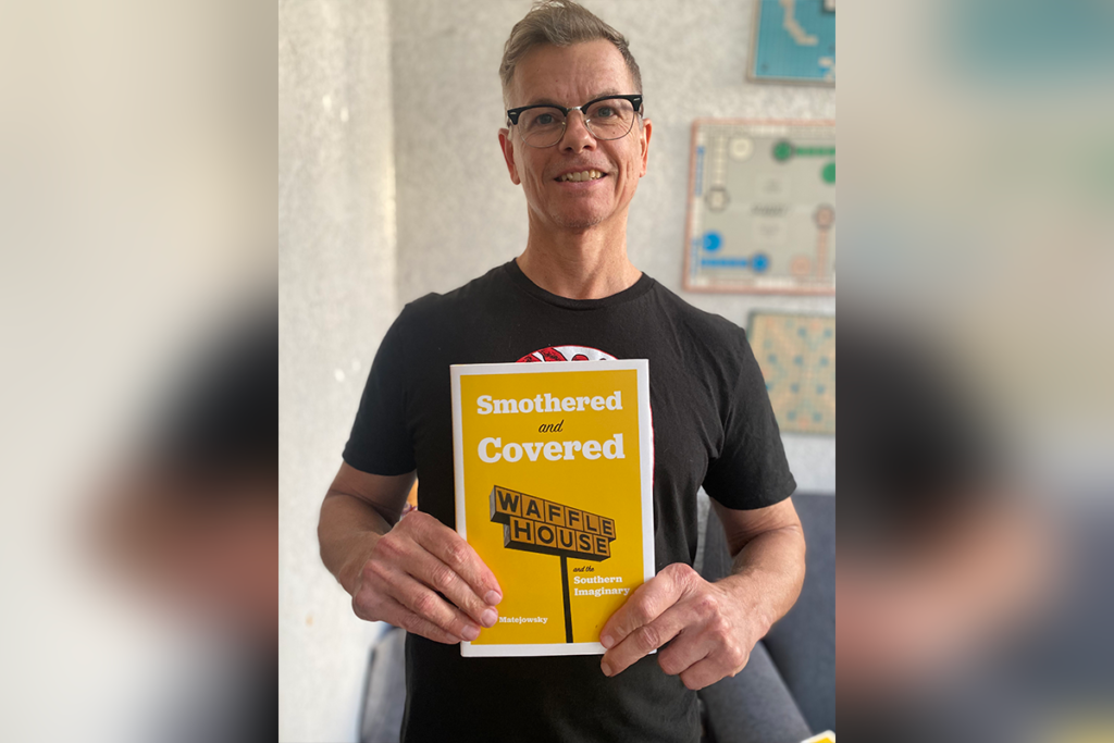 A man in glasses and a black shirt holds a yellow book titled “Smothered and Covered: A Waffle House Guide to Southern Eating.”.