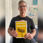 A man in glasses and a black shirt holds a yellow book titled “Smothered and Covered: A Waffle House Guide to Southern Eating.”.