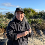 Smiling person in a black jacket and cap holding a small bird outdoors, with vegetation and blue sky in the background.