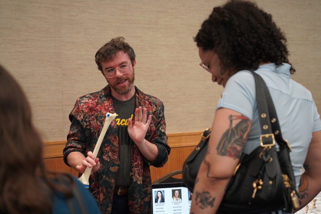 A man in a floral blazer holds a model human bone while speaking to two people.