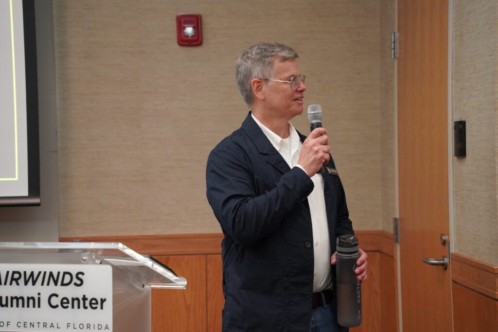 A man in a navy jacket speaks into a microphone while standing at a podium.