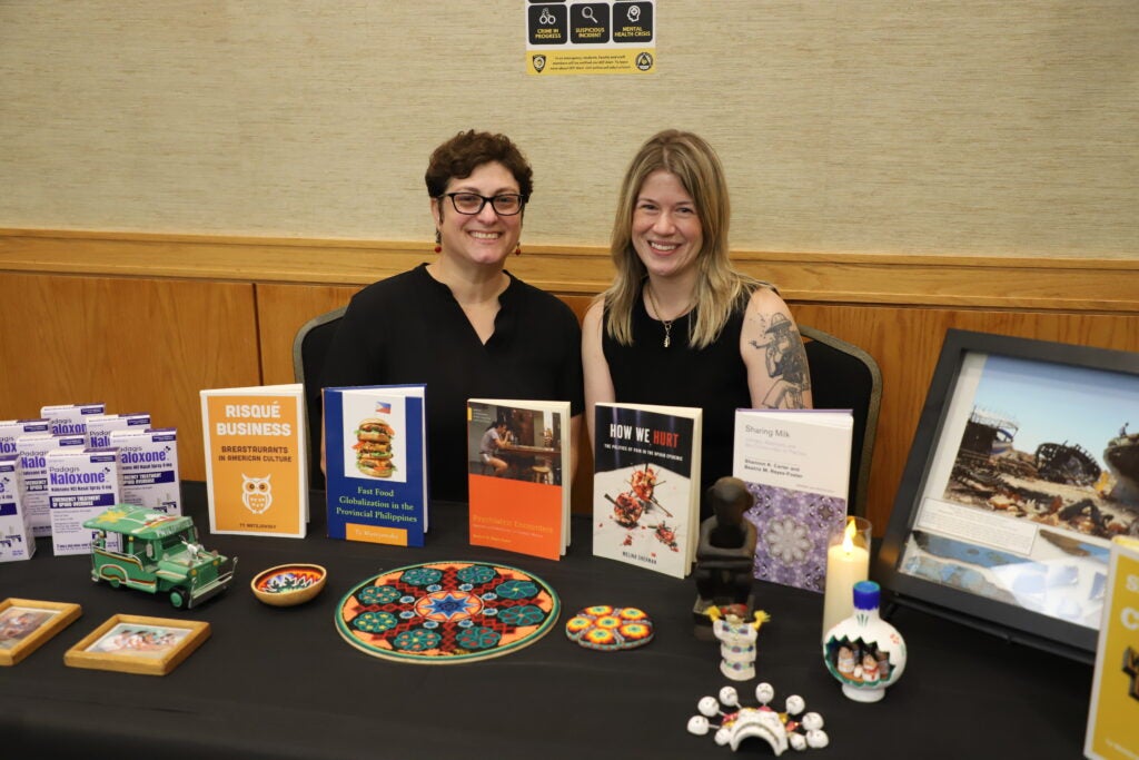 Two women smiling behind a table displaying several books, medicine boxes, and decorative objects.