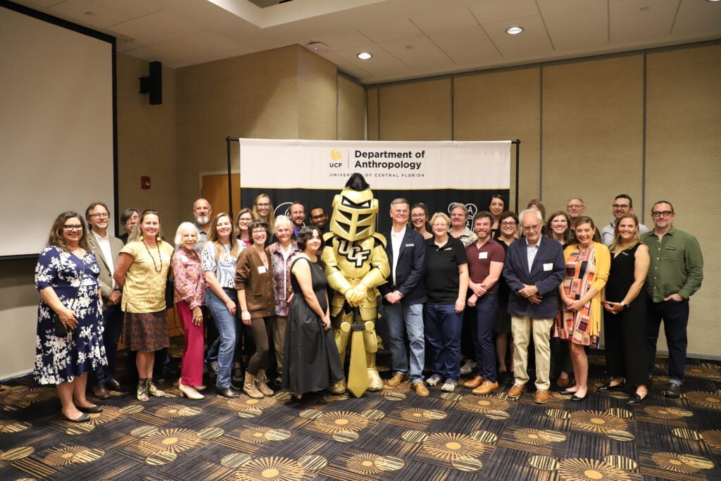A large group of faculty and staff from the UCF Department of Anthropology posing for a photo with the university mascot, Knightro, in front of a department banner.