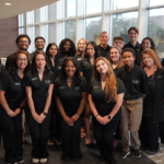 A group of students and staff in matching black polo shirts posing for a photo in an office building atrium.