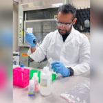 A scientist in a white lab coat and safety glasses uses a pipette to transfer liquid into a small vial in a laboratory.