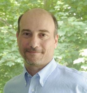 A man with short hair and a goatee is wearing a light blue collared shirt. He is standing in front of a background filled with green leaves.