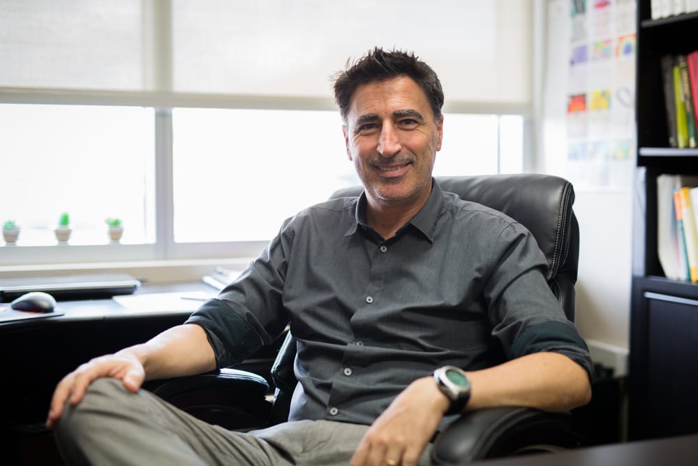 A man with dark hair and a grey shirt sitting in a black leather office chair, smiling at the camera in a well-lit office with shelves, books, and small plants near the window in the background.