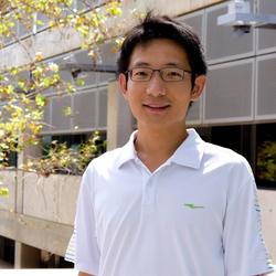 A person wearing glasses and a white polo shirt stands outside a building with large windows and leafy branches visible.