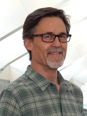 A man with glasses and a graying beard, wearing a green plaid shirt, stands indoors with a neutral expression.