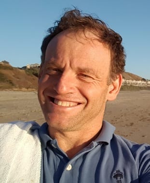 A man with short hair is smiling, wearing a blue polo shirt, and standing on a beach with hills and buildings in the background. A towel is draped over his left shoulder.