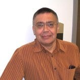 A man wearing glasses and a brown striped shirt stands indoors, leaning slightly to his right, with a neutral expression.