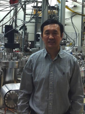 A man in a gray button-up shirt stands in front of complex laboratory equipment and wires.