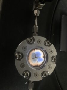 Round metallic chamber with several bolts and a glass window, showing a blue-violet plasma or glow inside, intersected by dark cross-shaped lines.