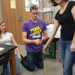 Three individuals are engaged in an activity inside a room. One person is using a tape measure, while the other two observe. The setting appears to be educational or workshop-related.