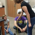 Three people are in a room with lab equipment. One person stands holding a measuring tape to the foot of a seated individual, while another person sits nearby observing.