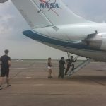 People boarding a NASA aircraft via stairs at the rear under cloudy skies, with individuals walking toward the plane on the tarmac.