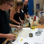 Three people assembling cylindrical devices on a workbench in a lab setting. Various tools and components are spread out on the table along with a chart.