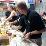 A group of people working on a project at a table in a workshop. Various tools, cups, and materials are scattered on the table. An American flag and some sports pennants hang in the background.