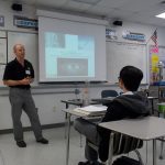 A man stands in front of a classroom delivering a presentation. The whiteboard behind him has notes and the projector screen displays a slide with various images and text. Students sit at desks listening.