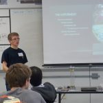 A person presents a scientific experiment in front of a classroom. The projected slide lists topics such as planetary formation and Saturn's shepherd moons. A whiteboard and seated audience are visible.