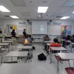 A classroom with students seated at desks facing a projector screen. Three individuals are standing in front of the class, apparently giving a presentation. The room is brightly lit with ceiling lights.