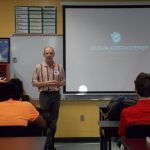 A teacher gives a presentation titled "Collisional Accretion Experiment" to a classroom of seated students. The classroom has a physics poster, bookshelves, and a whiteboard with the projection.