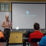 A man gives a presentation on a "Collisional Accretion Experiment" in a classroom. He stands in front of a projector screen, while students sit at desks listening.