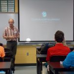 A man giving a presentation titled "Collisional Accretion Experiment" to a classroom of students seated at desks facing the projector screen.