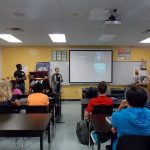 A classroom with students seated facing three presenters in front of a screen displaying a presentation. Classroom posters and lab equipment are visible on shelves and counters at the back.