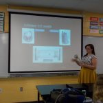 A woman stands in front of a projection screen labeled "EXPERIMENT TEST CHAMBER," holding a cylindrical object and explaining diagrams to an audience in a classroom.